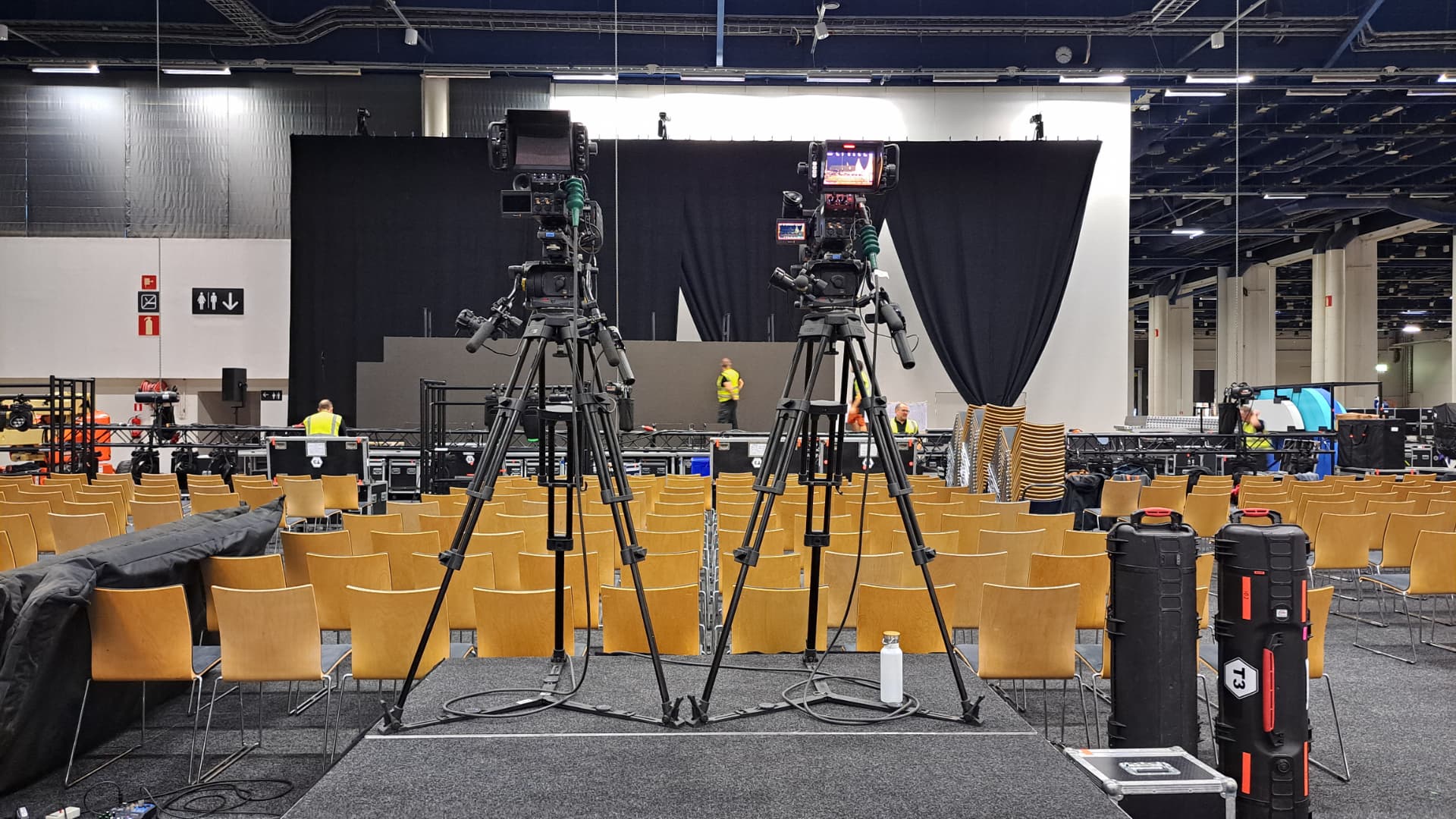Two broadcast cameras on tripods facing an empty stage in an event hall